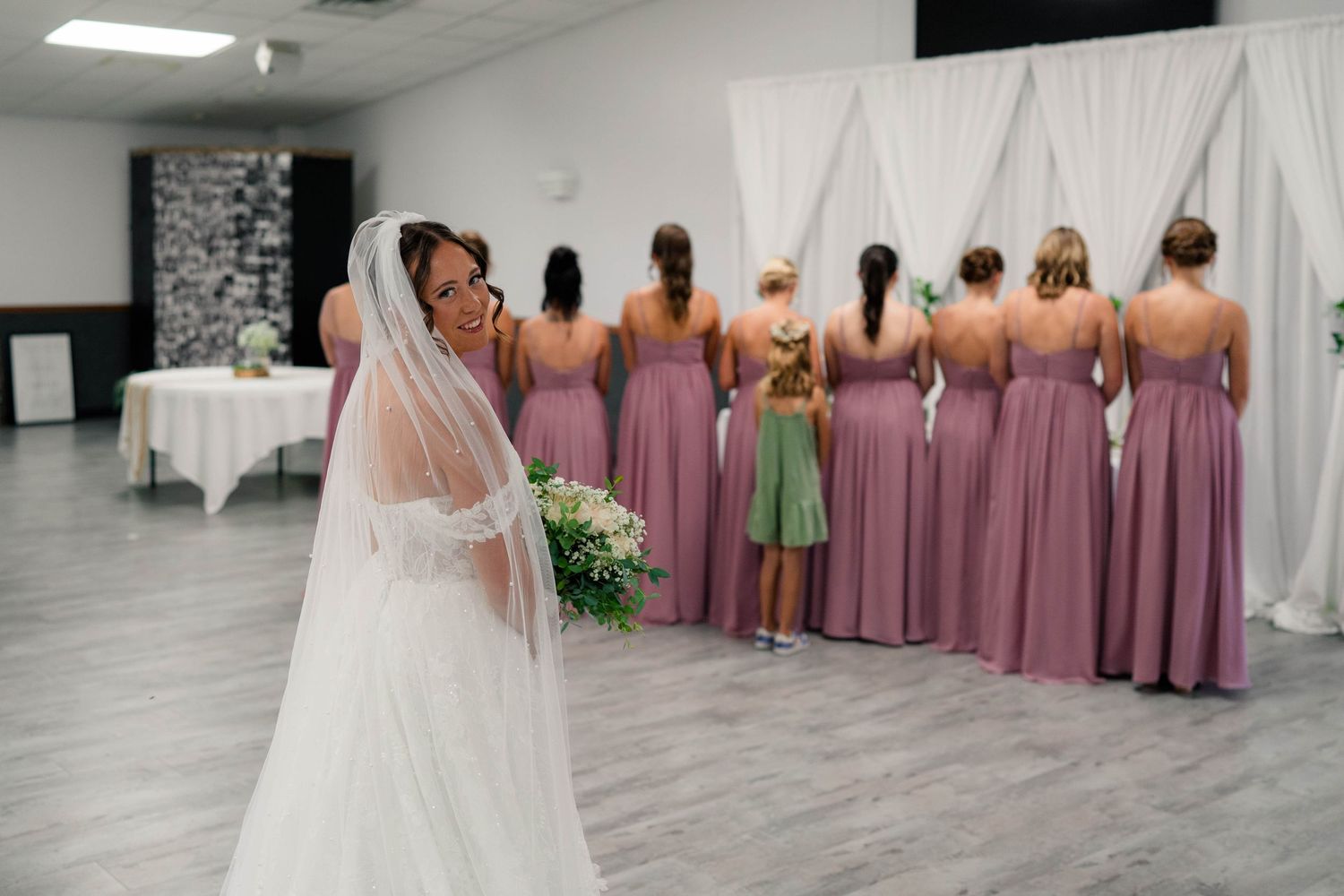 Bride in veil holding bouquet stands behind bridesmaids in mauve gowns lined up at ceremony backdrop.