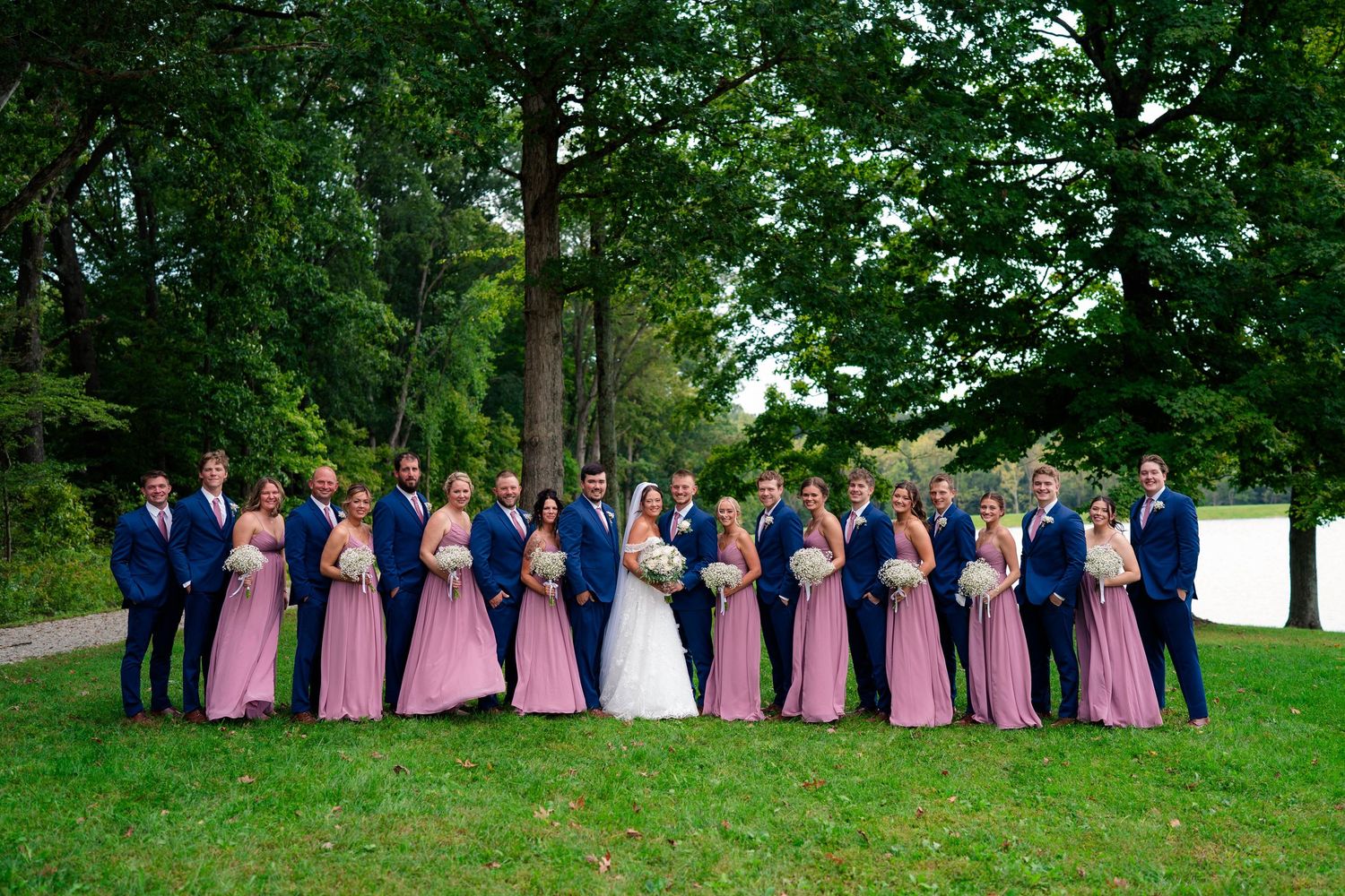 Large wedding party in navy suits and dusty rose gowns posing under tree by lake.