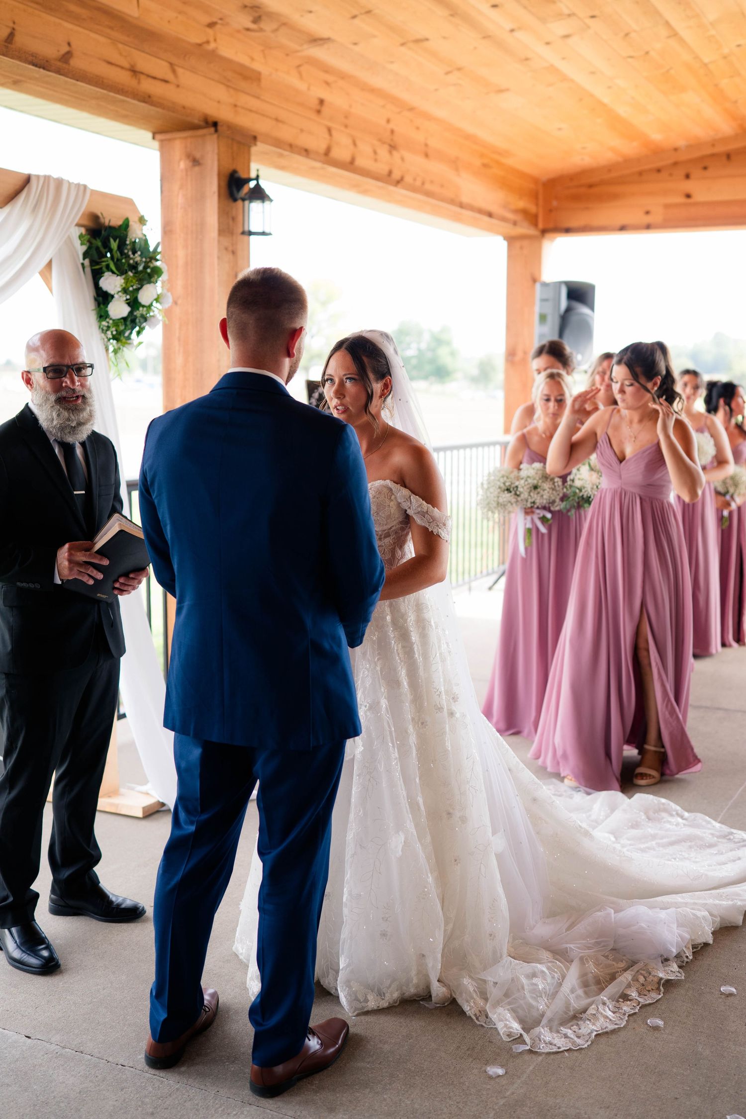 Bride and groom exchange vows under a wooden pavilion as bridesmaids in dusty rose gowns look on.