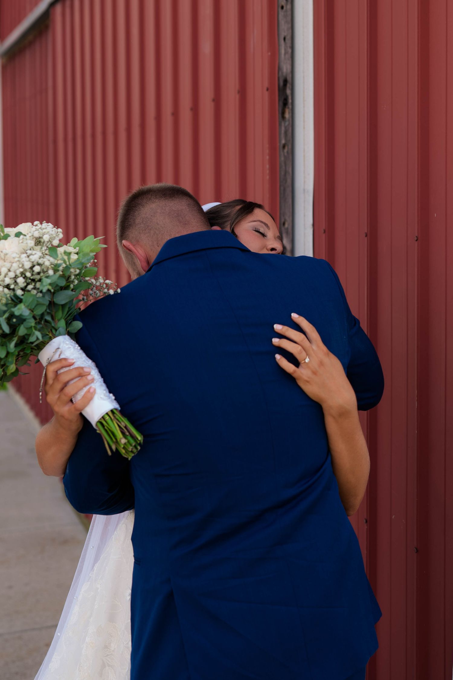 Bride and groom share an intimate embrace against a red barn wall, bride holding a white floral bouquet.