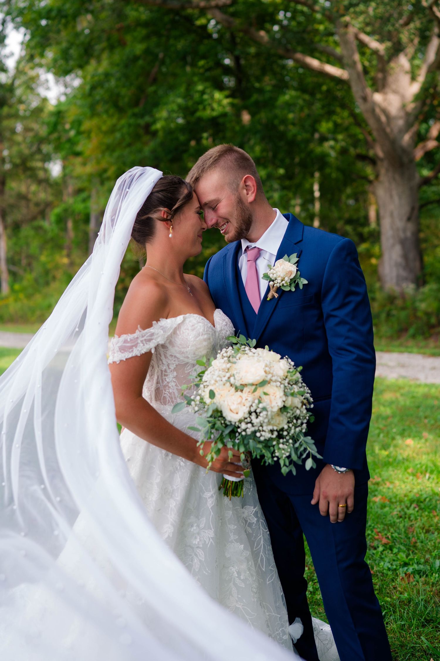 Bride and groom touching foreheads romantically outdoors, bride wearing lace off-shoulder gown with flowing veil.