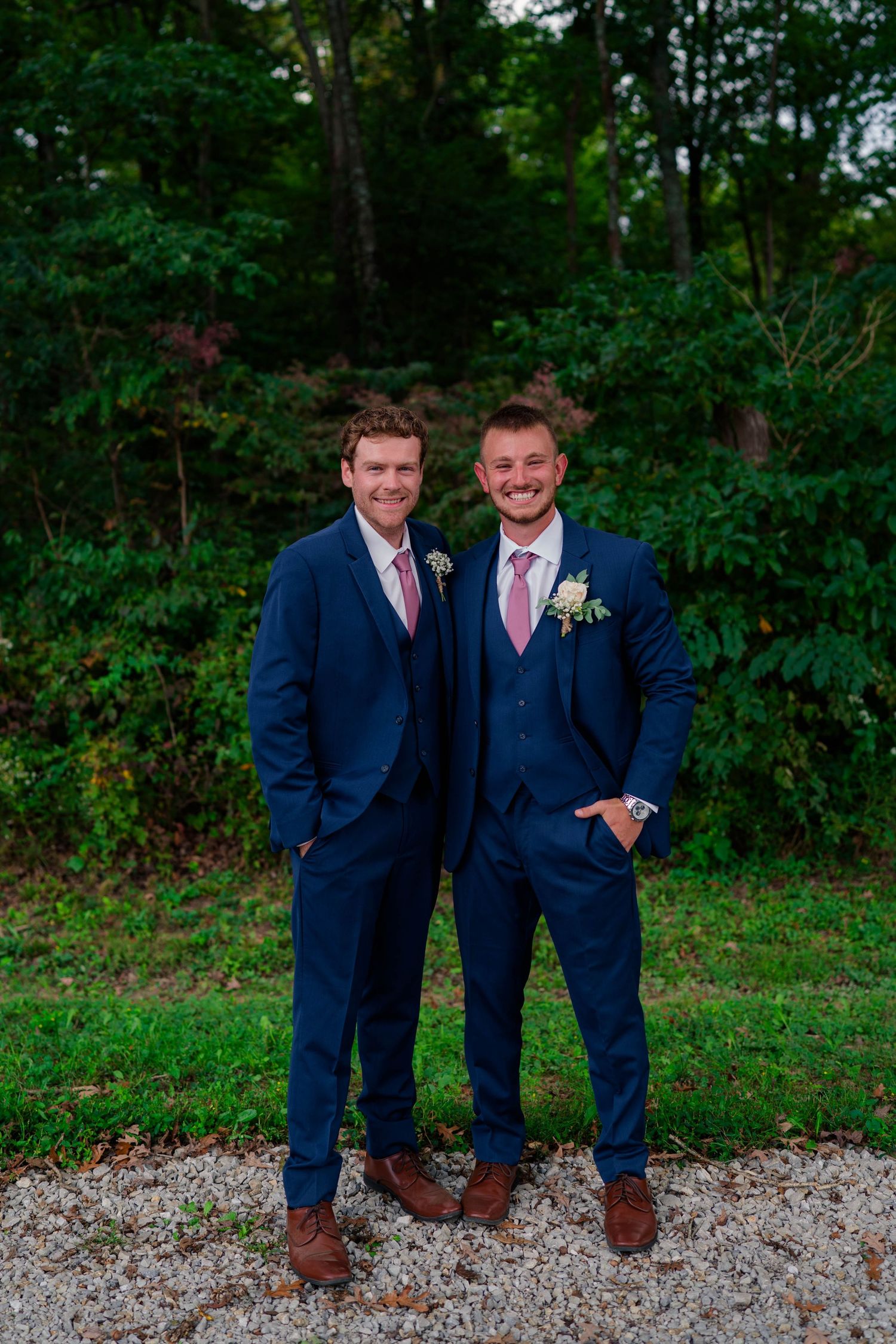 Two smiling groomsmen in navy blue suits with pink ties posing outdoors.