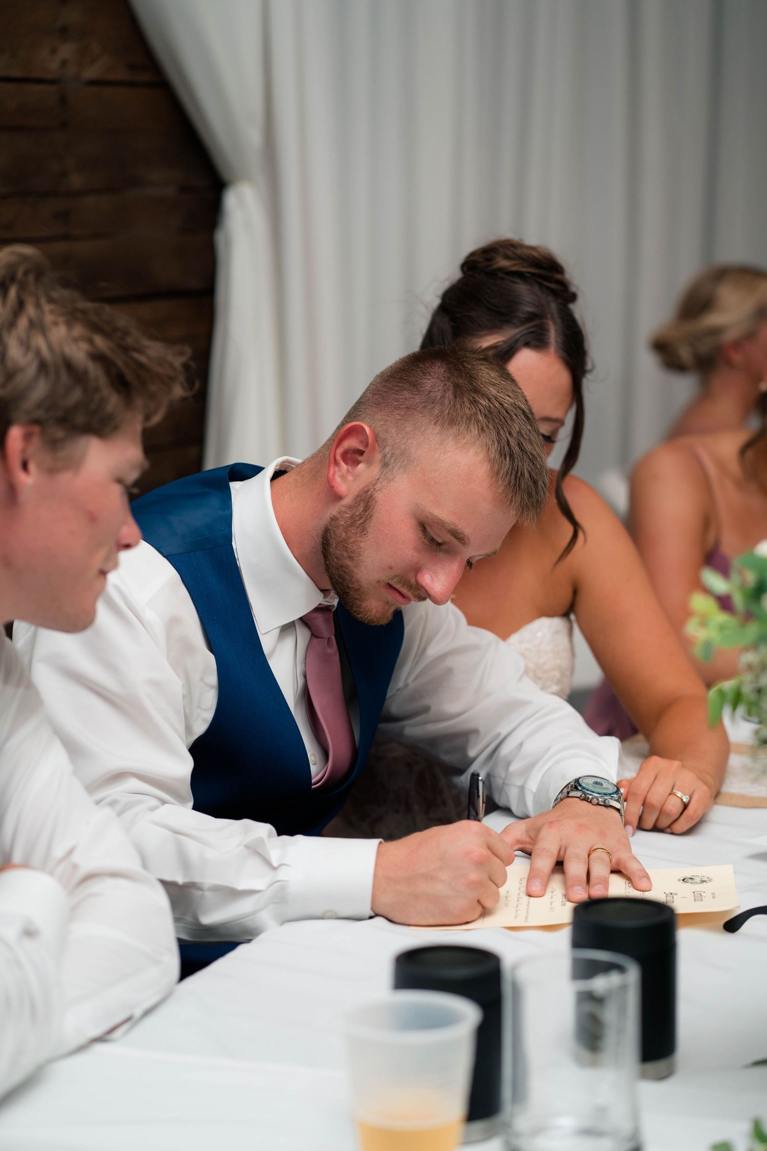 Groomsman in navy vest and pink tie writing on paper at the wedding head table with bride visible in background.