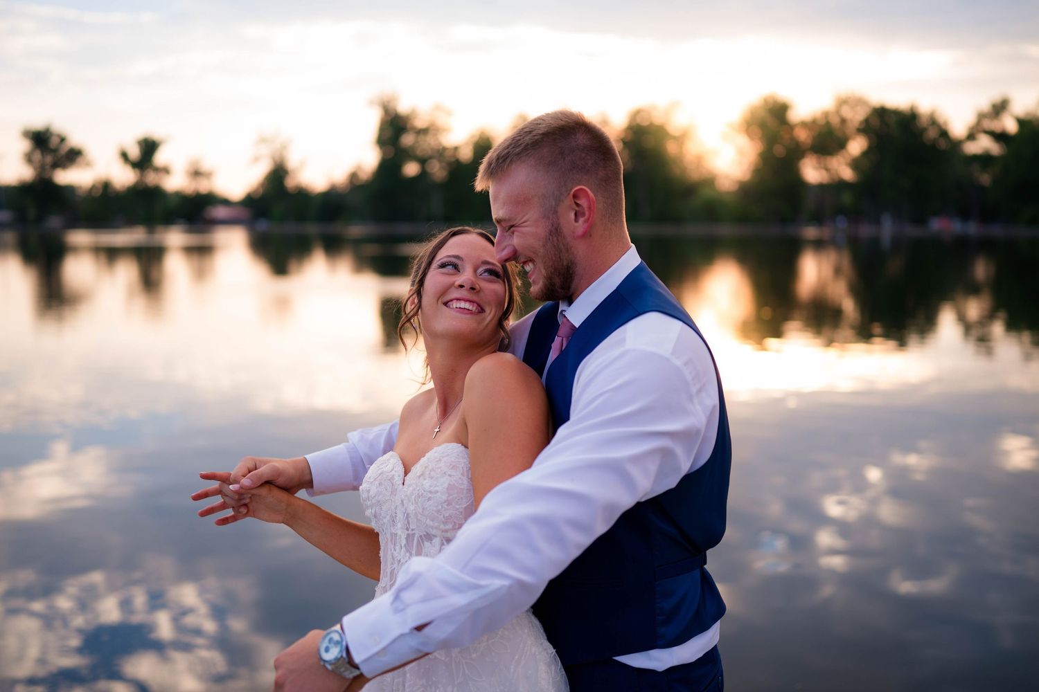 Bride and groom share a romantic embrace at sunset by a reflective lake, smiling joyfully on their wedding day.