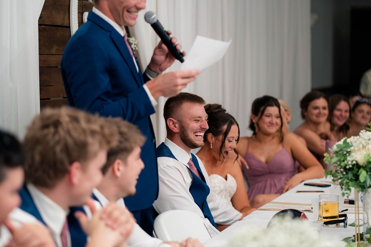 Wedding reception speech moment with groom laughing at head table while guests smile and clap in elegant white venue.