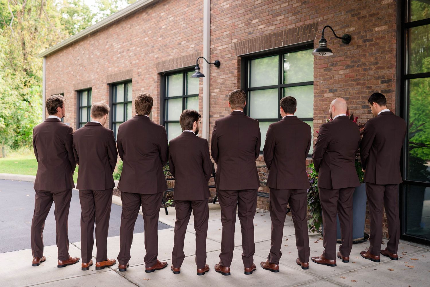 Groomsmen in brown suits stand in a row facing brick building exterior at wedding venue.