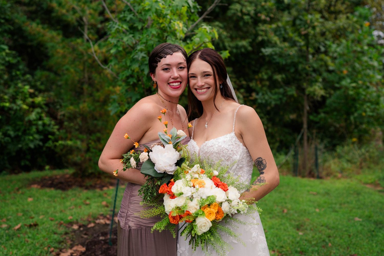 Smiling bride with orange and white bouquet poses with bridesmaid in taupe dress outdoors.