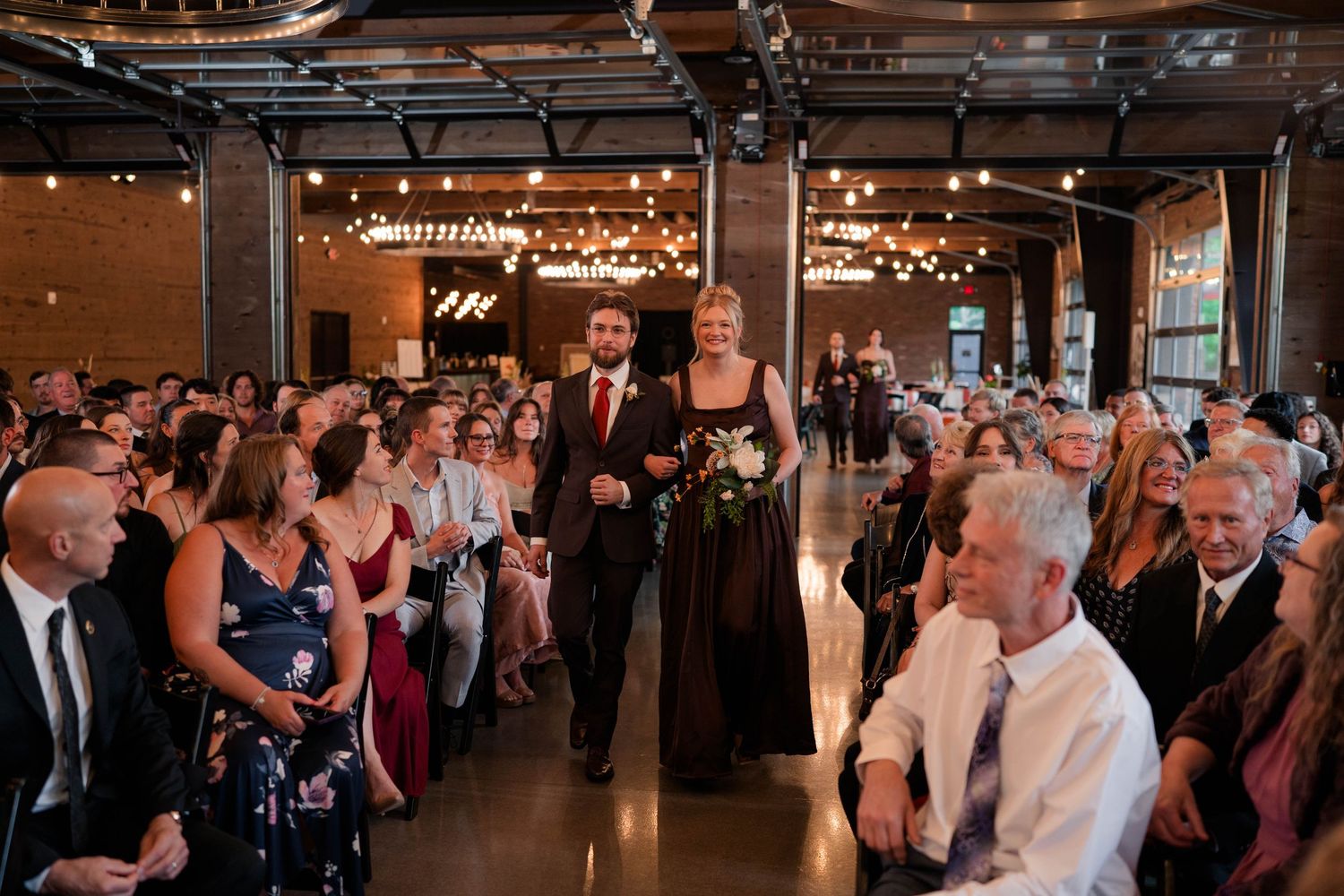 Bridesmaid in black gown walks down the aisle with escort at an elegant indoor wedding ceremony.