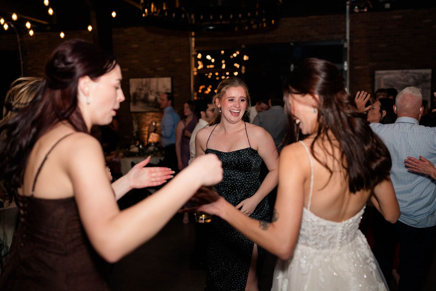 Bridesmaids in black dresses dance joyfully with bride at wedding reception under string lights.