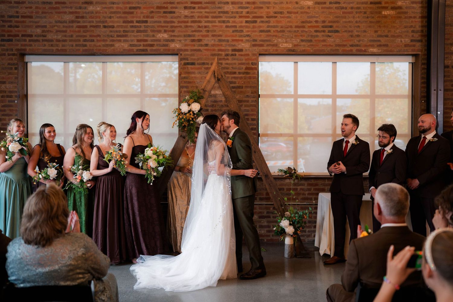 Bride and groom share their first kiss at the altar surrounded by bridesmaids and groomsmen in a brick venue.