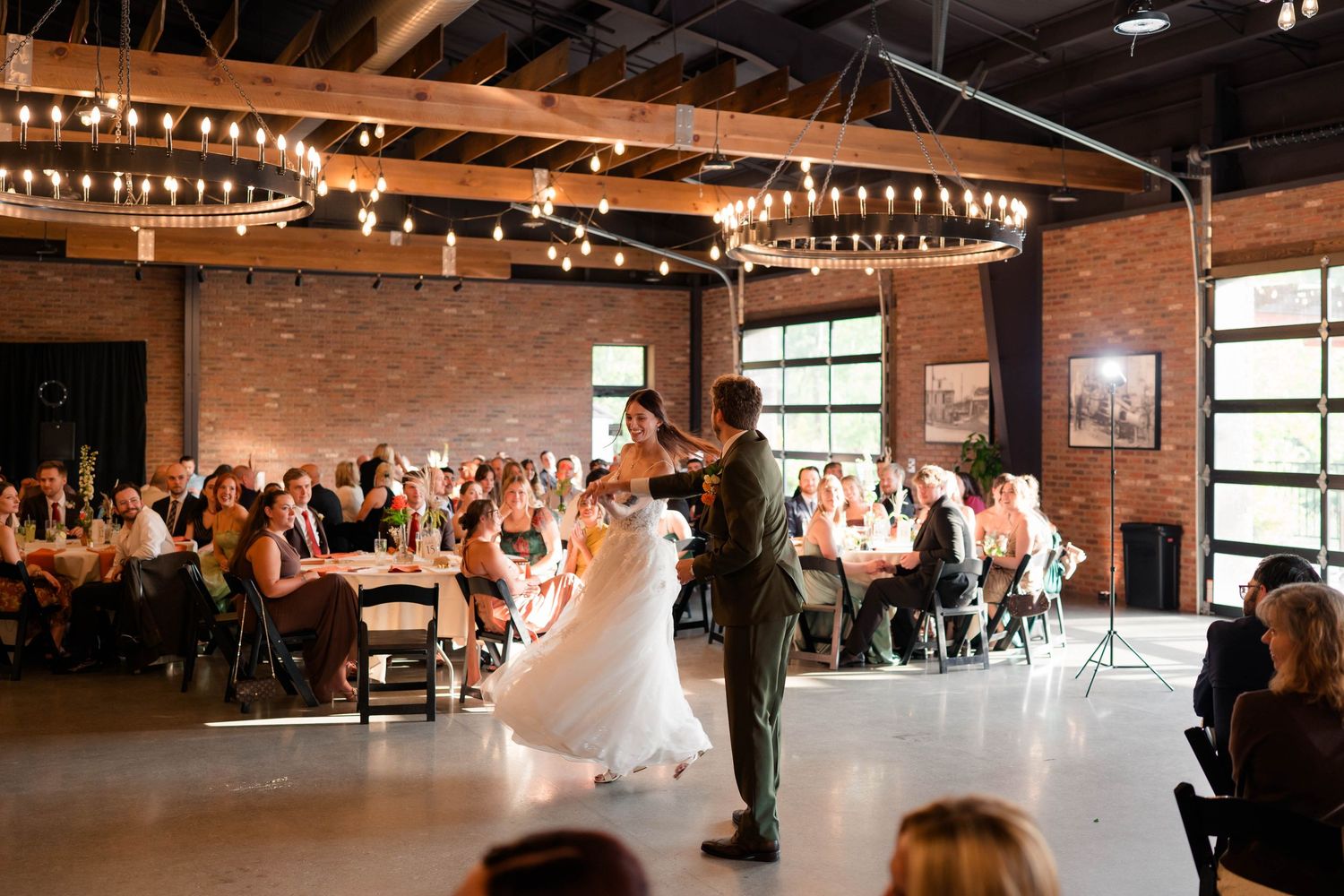 Wedding couple shares first dance as guests watch in warmly lit industrial brick reception hall.