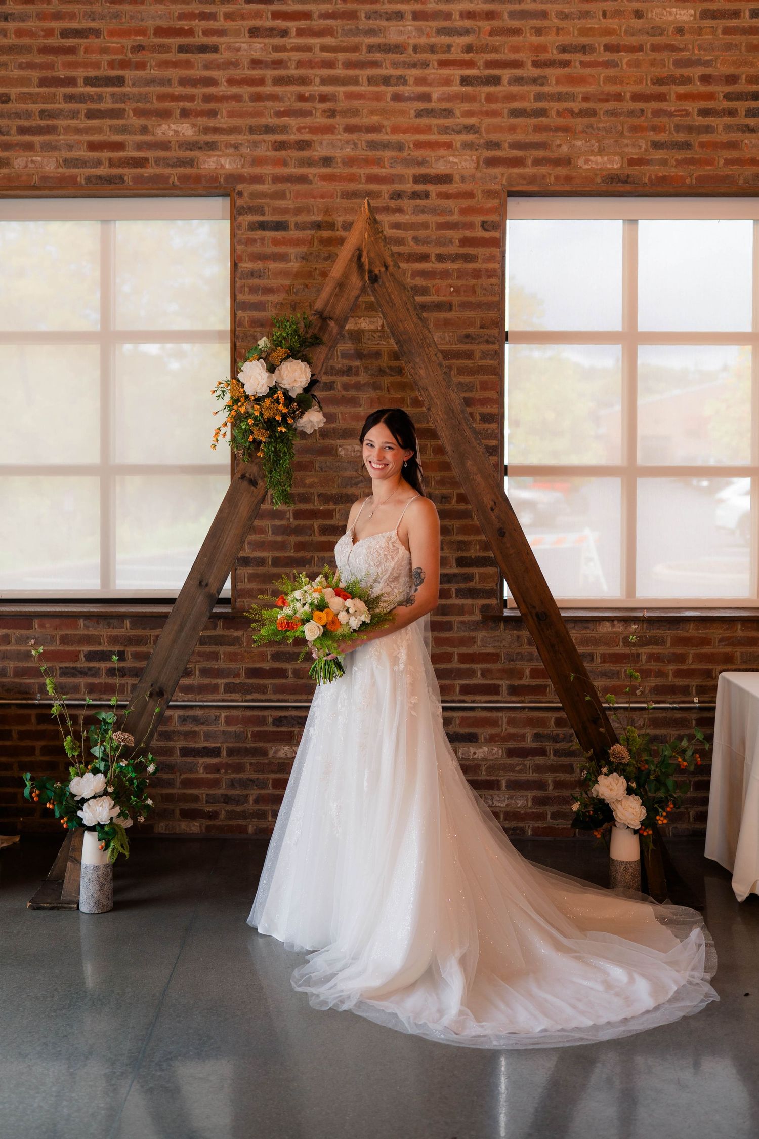 Bride in lace gown holds vibrant bouquet standing before a wooden triangular arch with florals in brick venue.