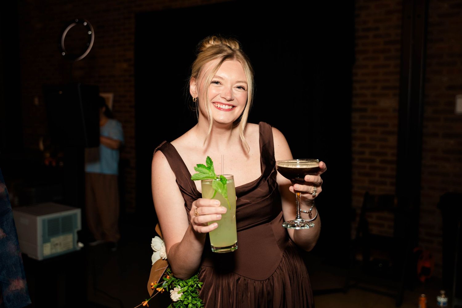 Smiling bridesmaid in brown dress holds green cocktail and martini glass at wedding reception.