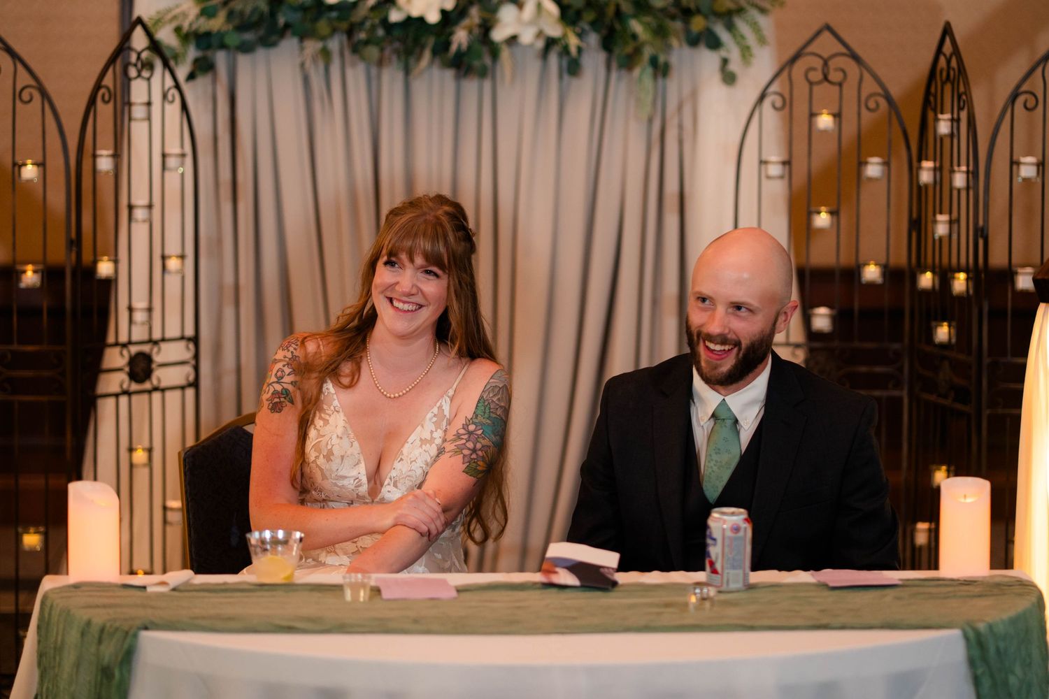 Bride and groom smiling at sweetheart table during candlelit wedding reception.