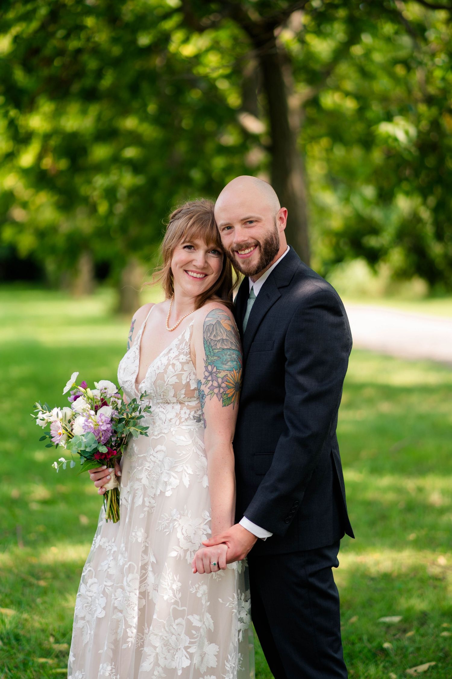 A smiling bride and bald groom pose together in a lush green park, bride holding a floral bouquet.