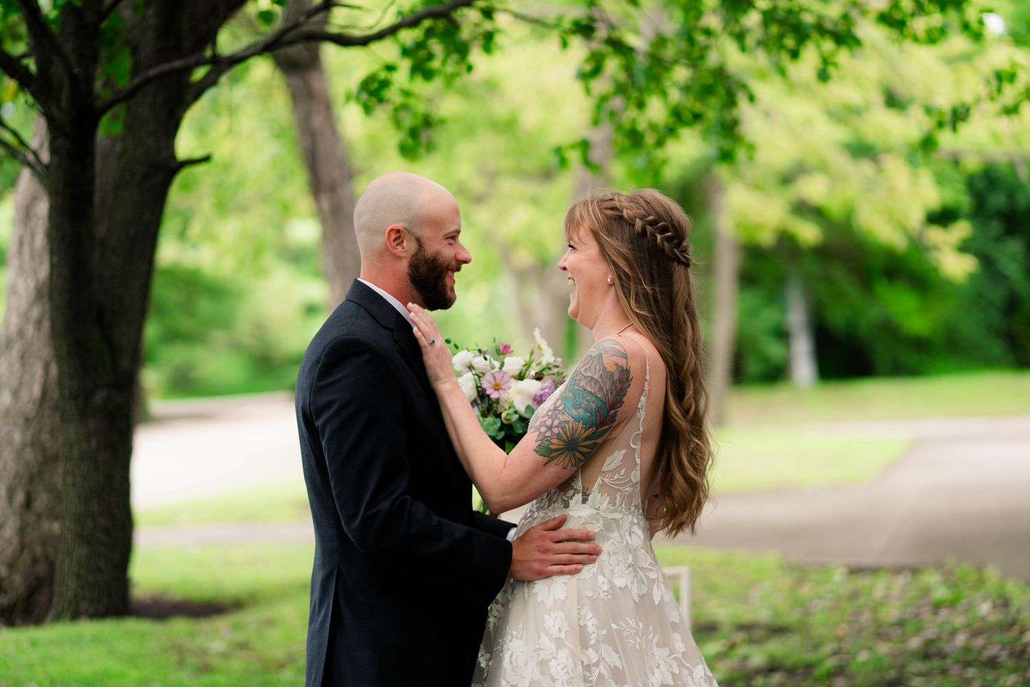 Bride and groom share a tender moment under green trees during outdoor wedding.