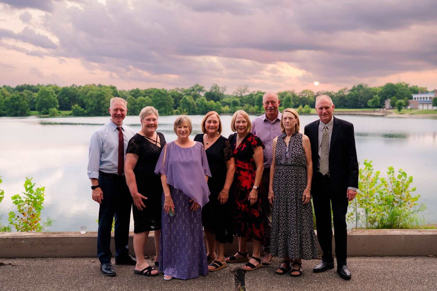 Group of eight wedding guests pose together outdoors with dramatic sunset sky and lake in background.