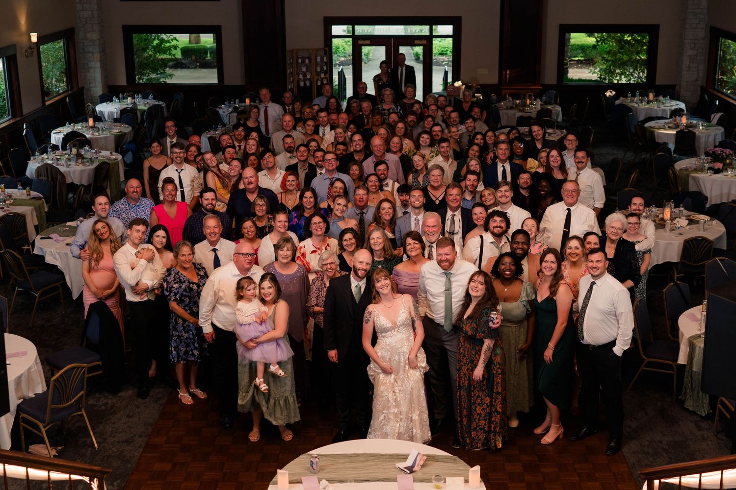 Large wedding group photo with bride, groom, and all guests gathered in reception hall.
