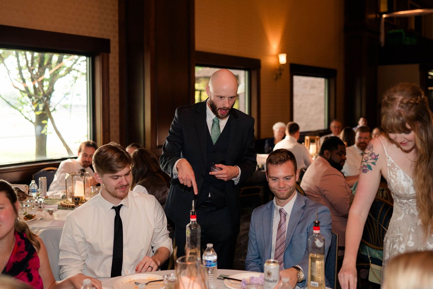 Bald groomsman in suit interacts with seated wedding guests at decorated reception dinner tables indoors.