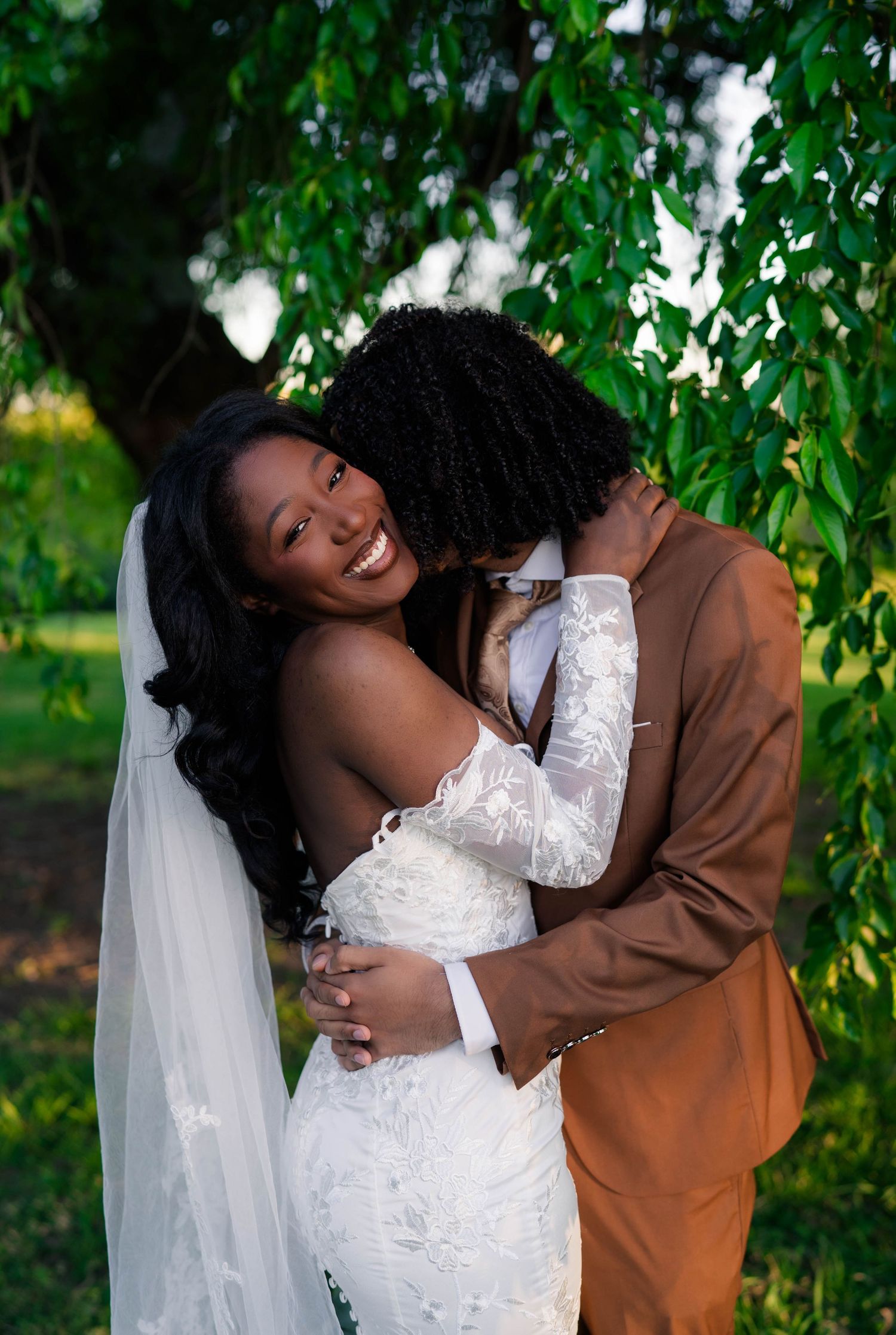 Smiling bride in white lace gown and veil embraces groom in brown suit under green willow tree.