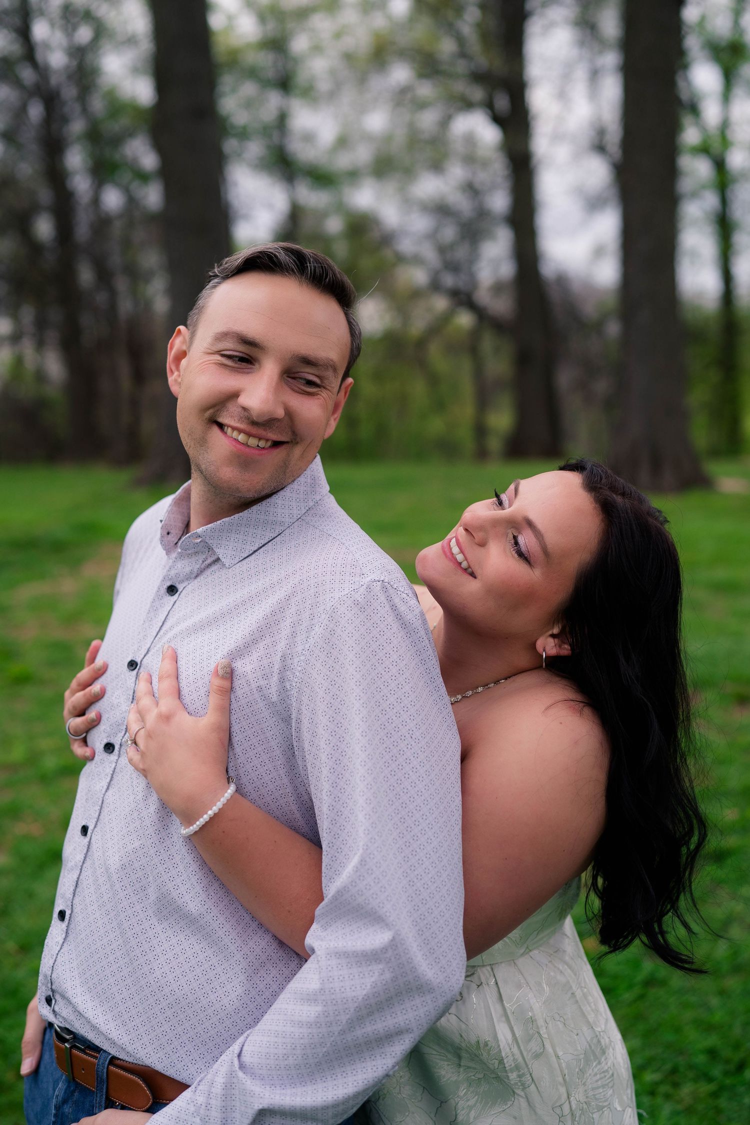 Smiling man embraced from behind by dark-haired woman in green dress among tall trees outdoors.