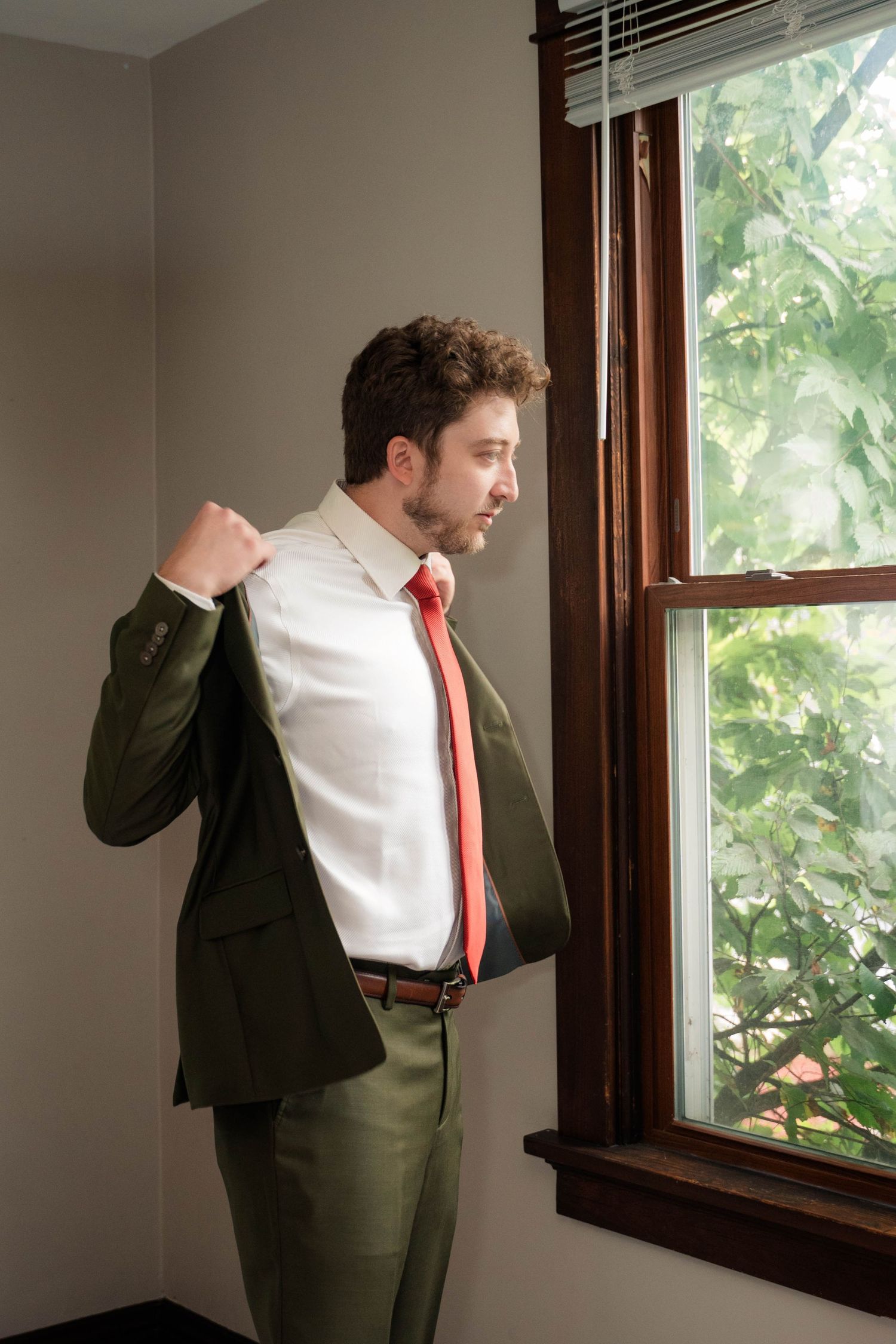 Groom in olive green suit and red tie adjusting jacket while looking out window.
