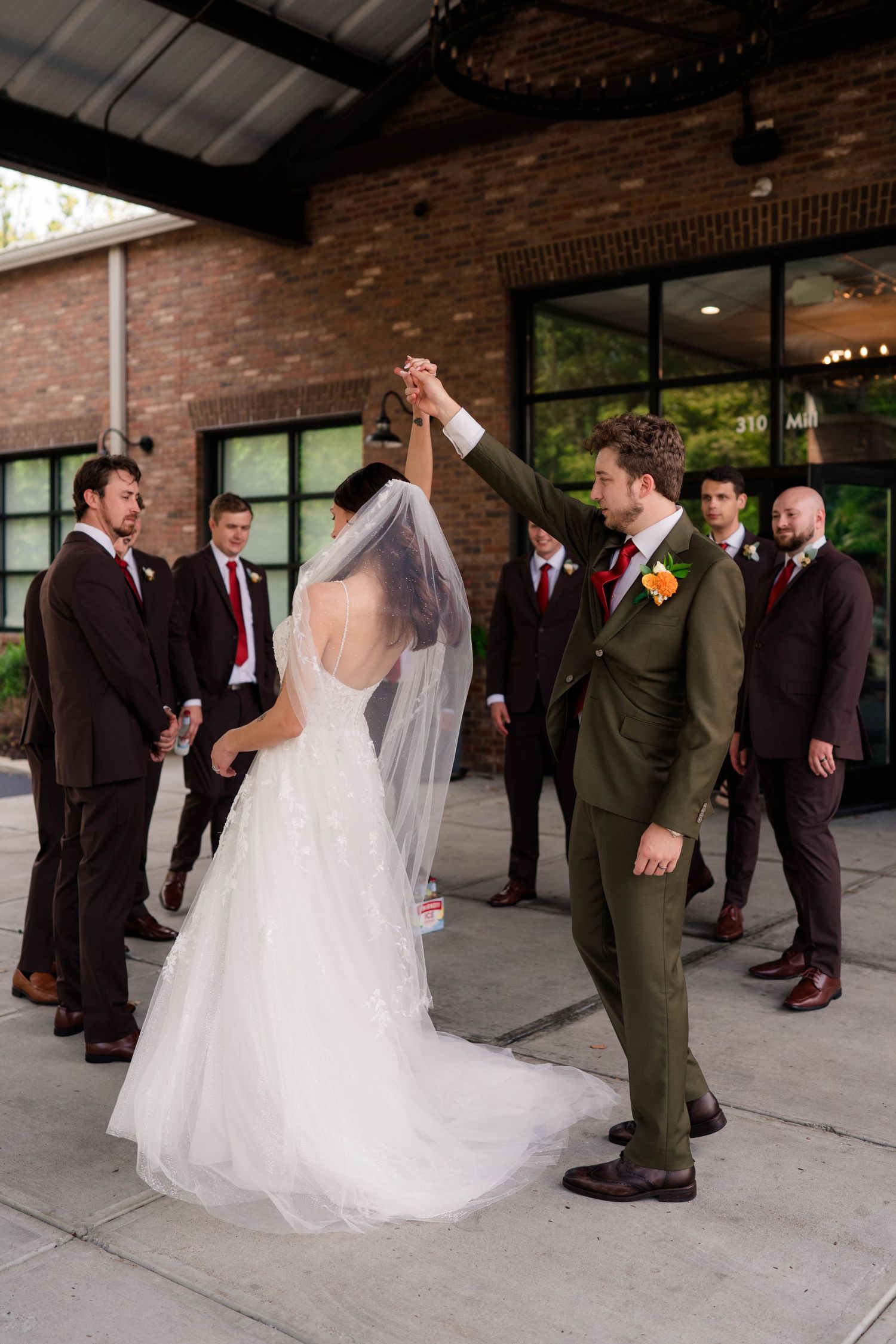 Groom spins bride in white gown while groomsmen in burgundy suits watch outside brick wedding venue.