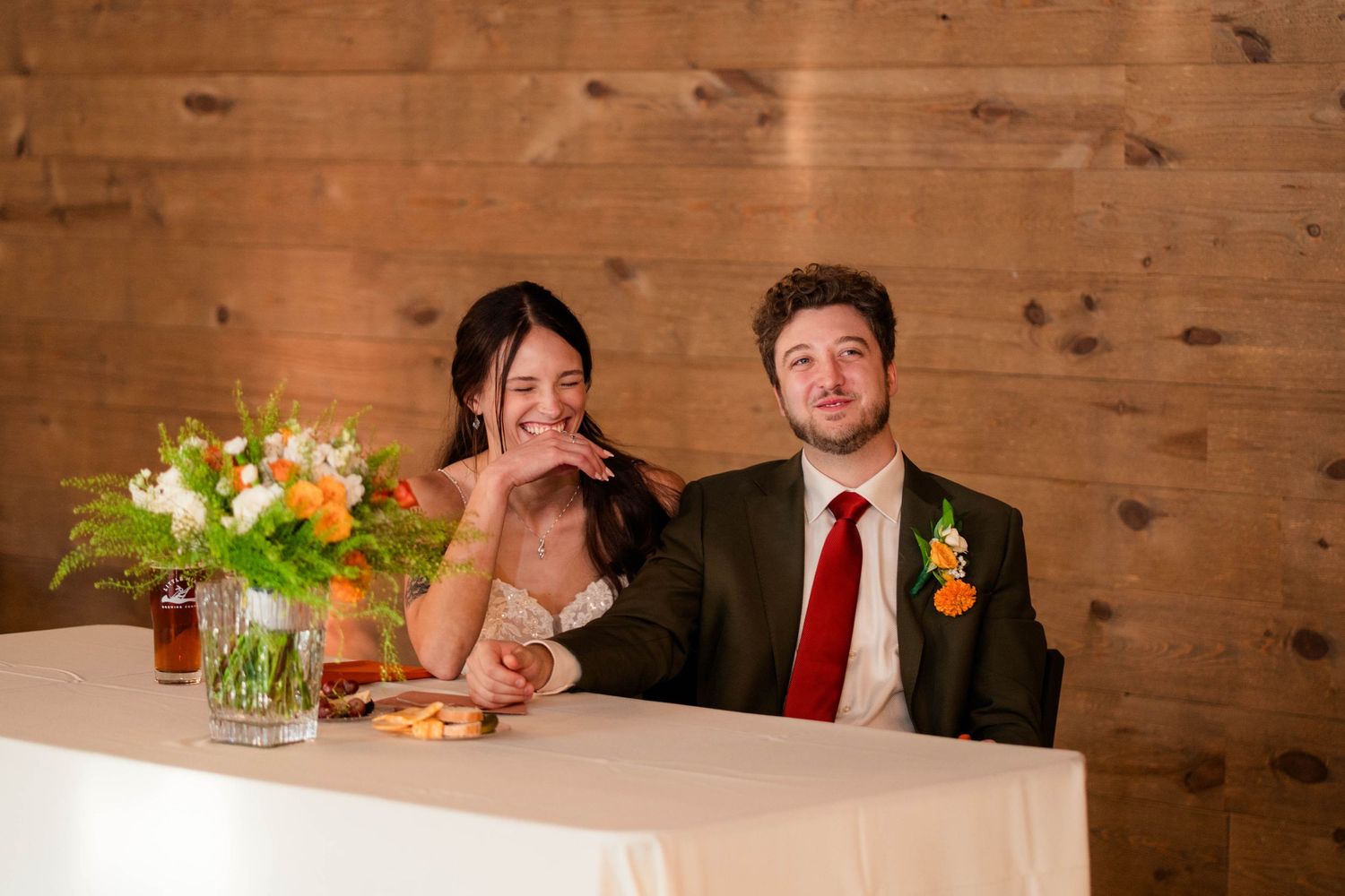 Bride and groom share a lighthearted moment at sweetheart table with colorful floral centerpiece.