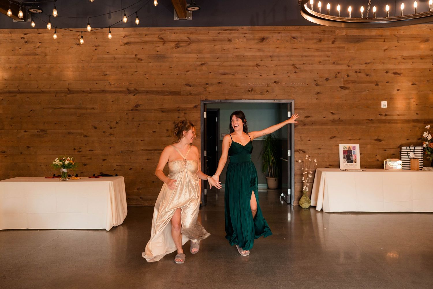 Two bridesmaids in green and gold dresses dancing together at a rustic wedding reception.