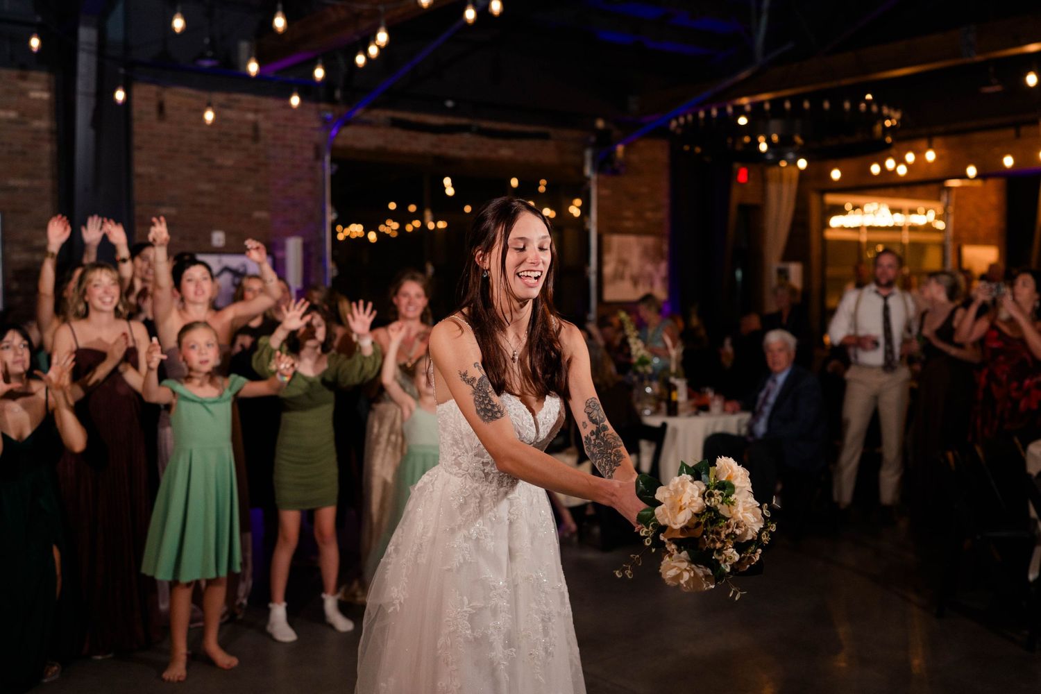 Bride smiling while tossing bouquet to excited guests at wedding reception in rustic venue.