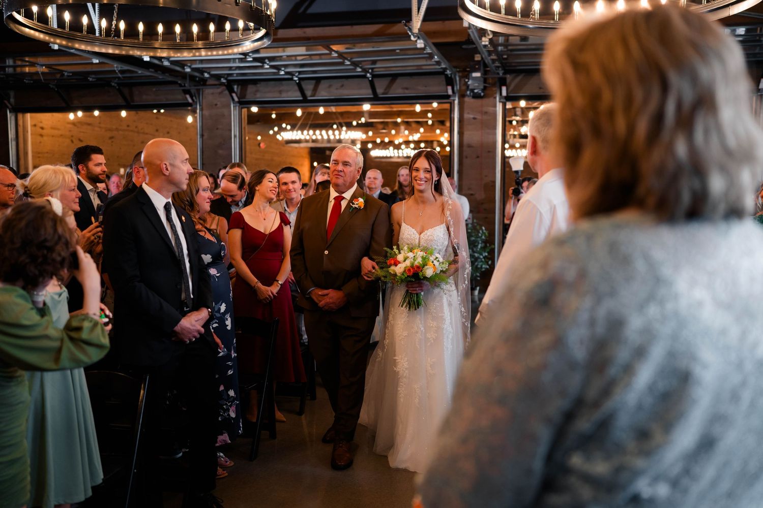 Bride walks down aisle with escort at indoor wedding ceremony with string lights.
