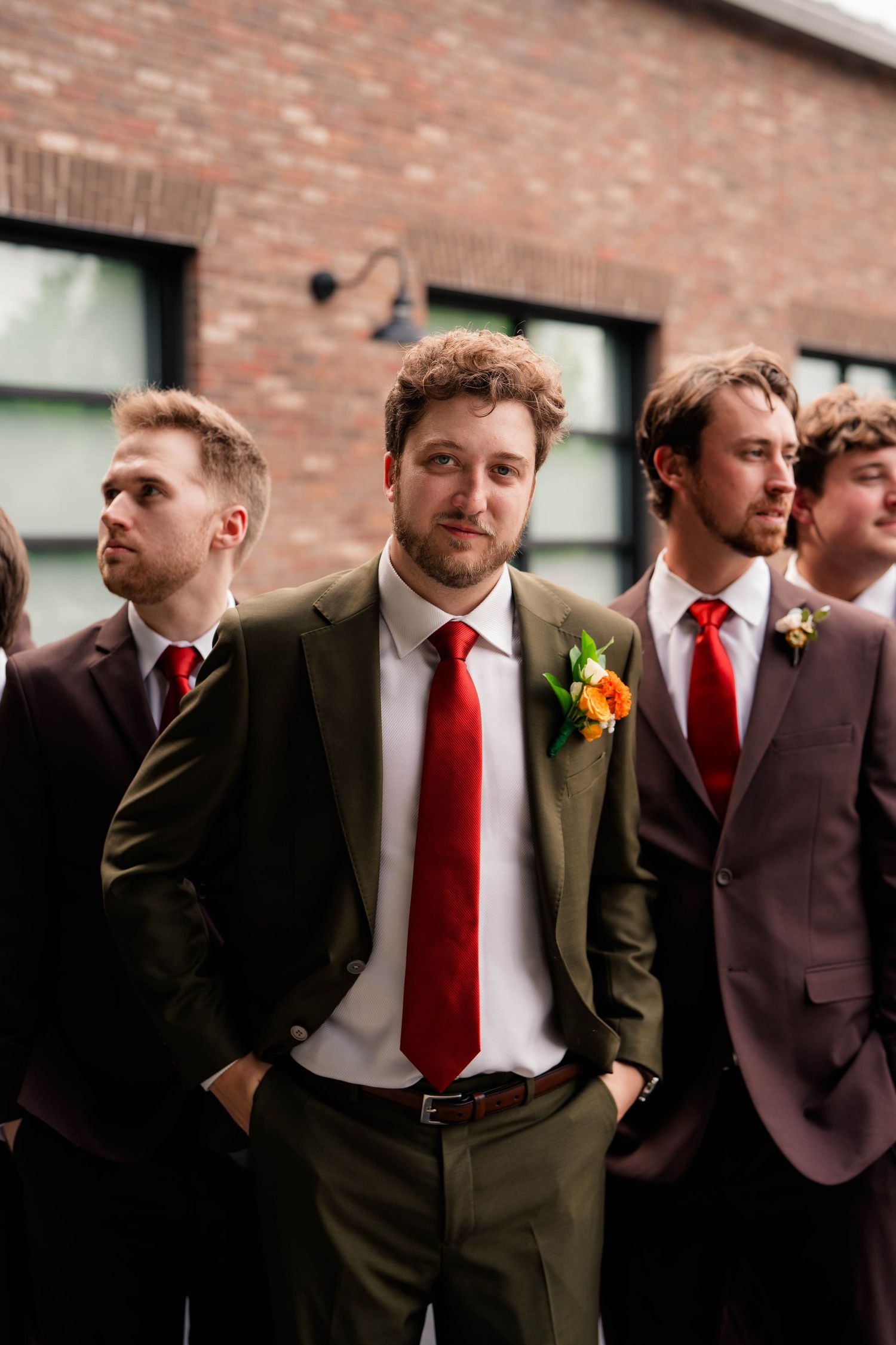 Groom in olive suit with red tie and boutonniere stands with groomsmen before wedding ceremony.