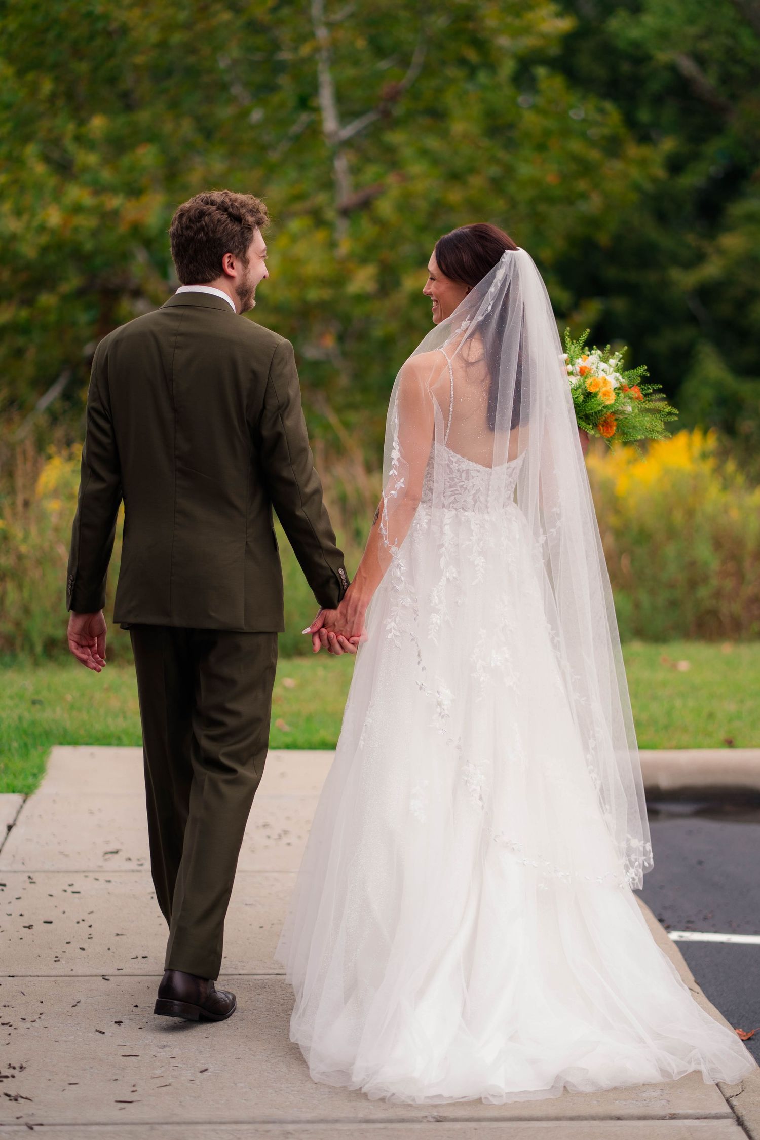 Bride and groom walk hand in hand outdoors, bride wearing lace gown with veil holding floral bouquet.