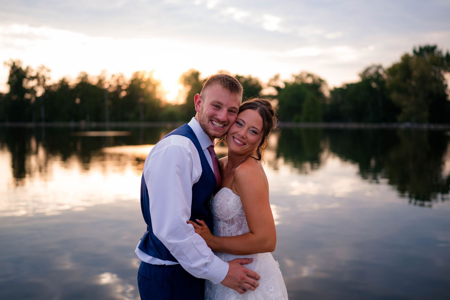 Happy wedding couple posing together by a serene lake at sunset, smiling joyfully in formal attire.
