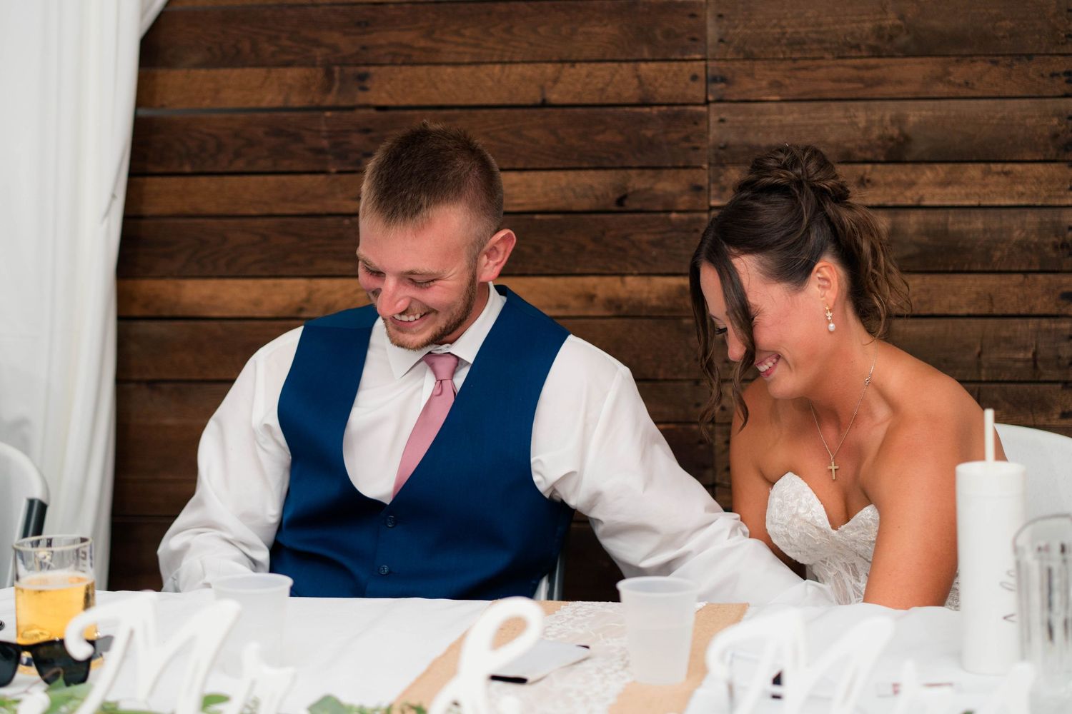 A smiling bride and groom sit together at their wedding reception table, dressed in bridal gown and navy vest.