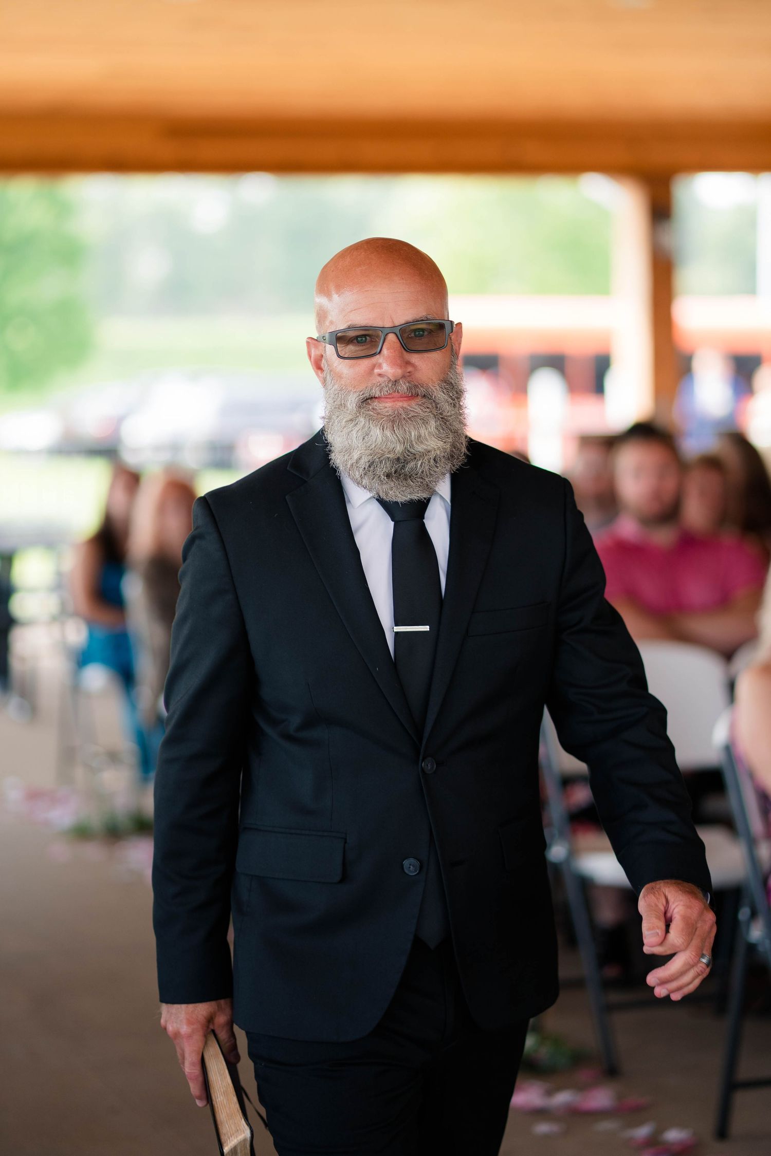 Bald man with gray beard in black suit and sunglasses walks down wedding ceremony aisle.