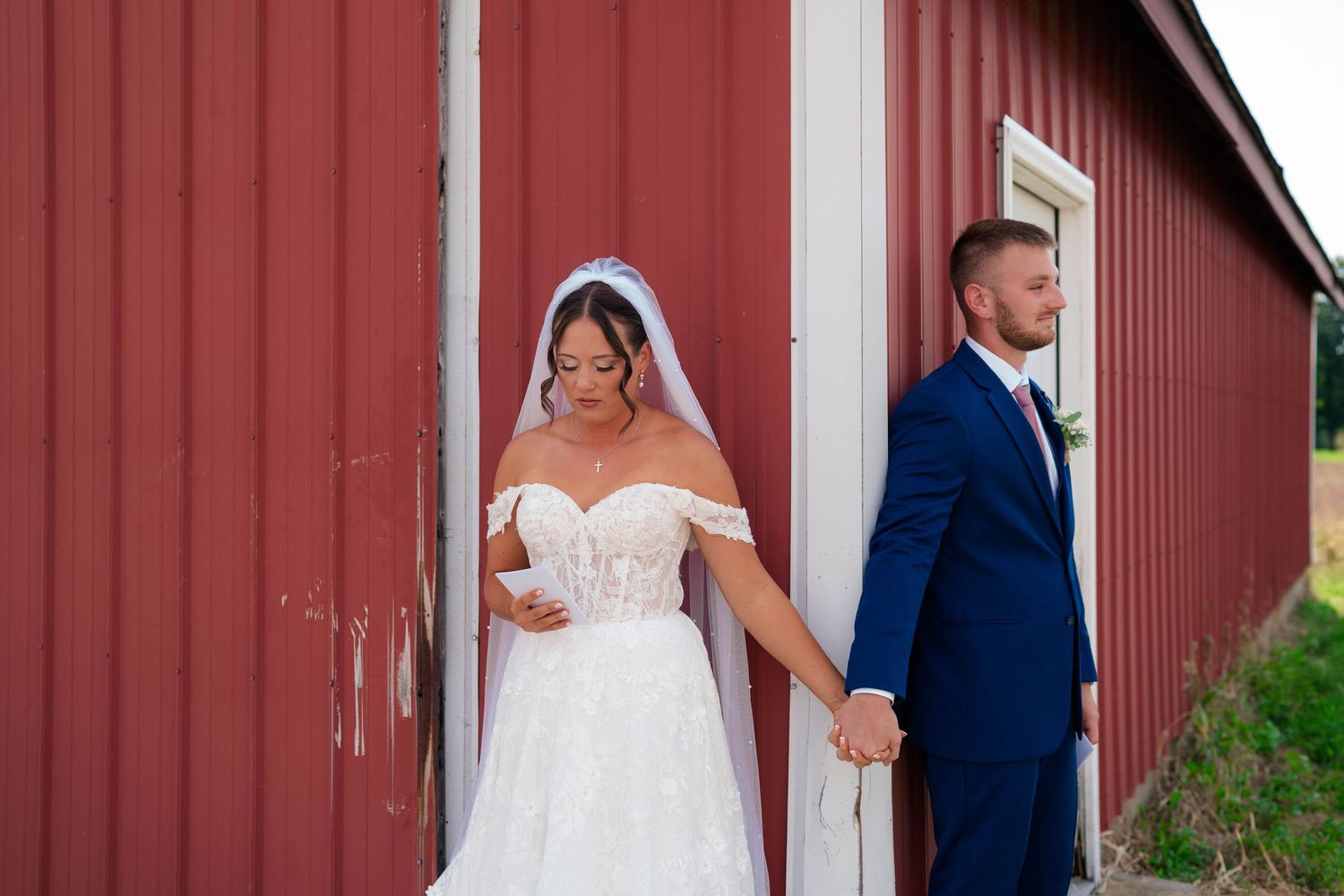 Bride in white off-shoulder gown and veil holds groom's hand in navy suit by a red barn.