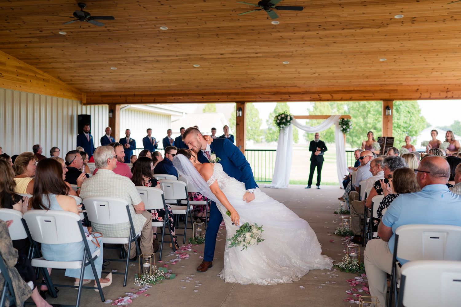 Groom dips bride for romantic kiss during wedding recessional with seated guests applauding.