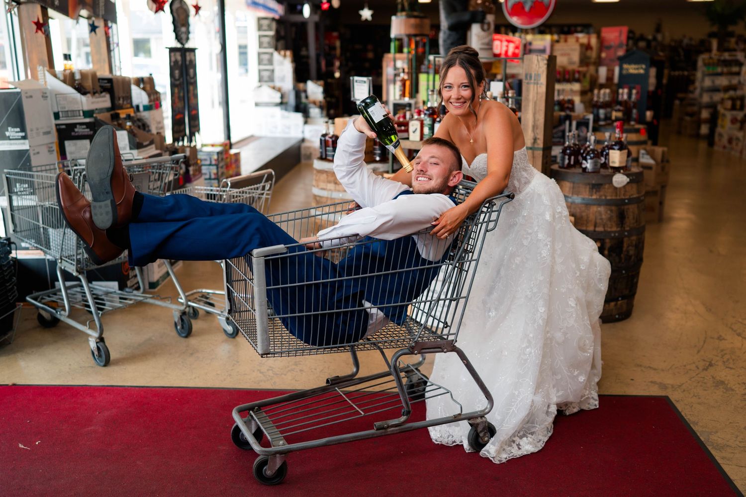 Bride pushes groom in shopping cart holding champagne bottle inside rustic store.