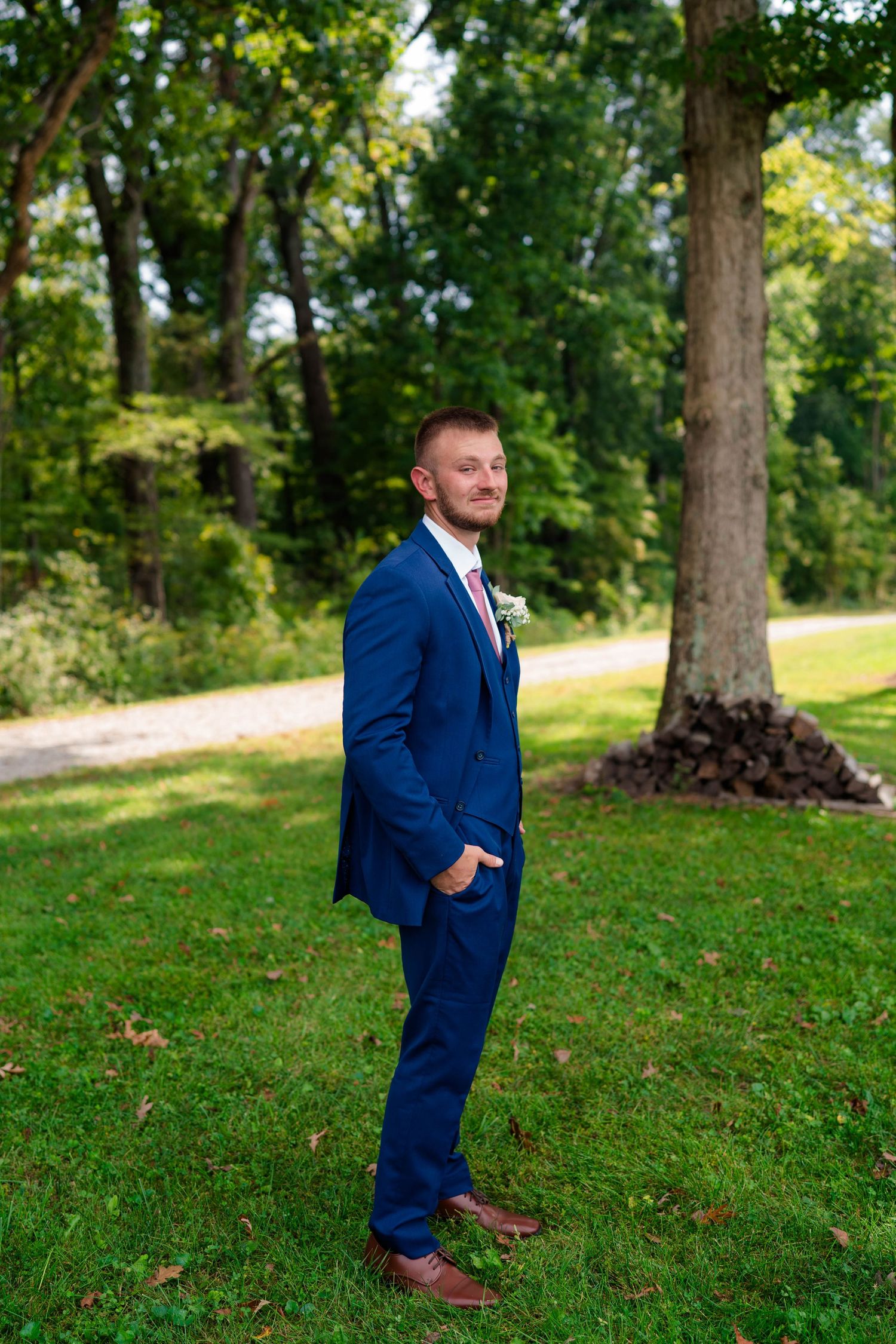 Groom in navy blue suit with boutonniere poses confidently on a green lawn by trees.