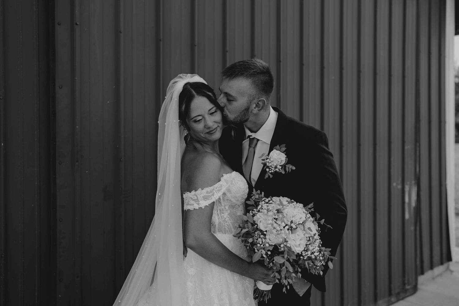 Black and white photo of a bride and groom sharing a tender moment, groom kissing bride's temple as she holds a bouquet.