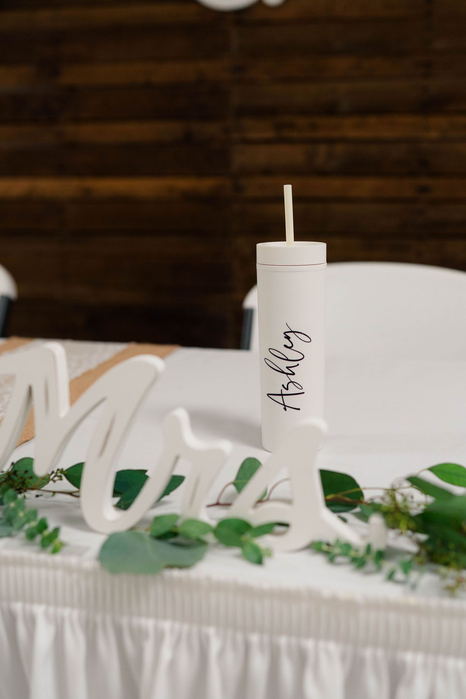 Personalized white tumbler with name Ashley sits beside Mrs. wooden sign decorated with greenery at wedding sweetheart table.