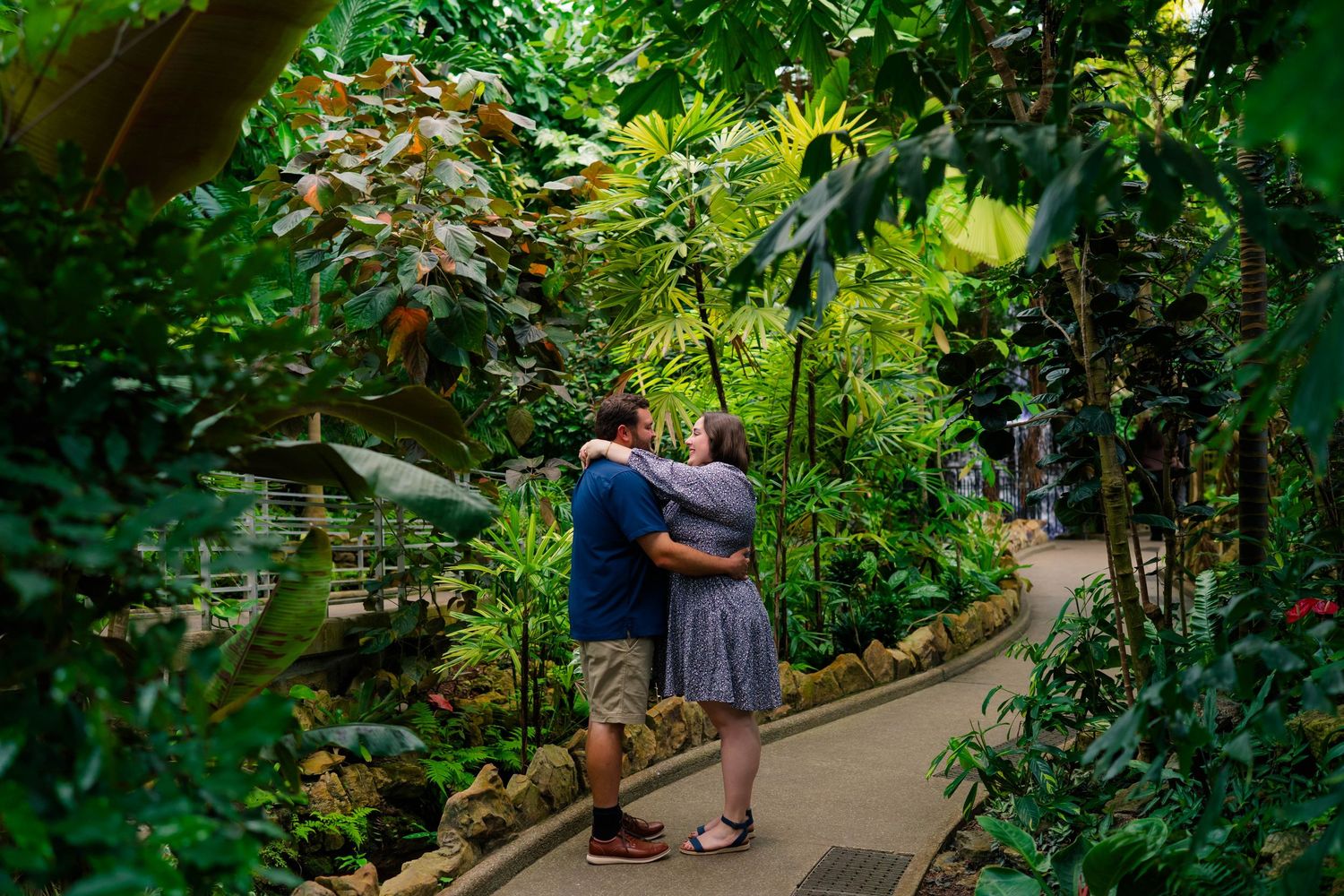 Engaged couple hugging on a garden path surrounded by tropical plants in a greenhouse.