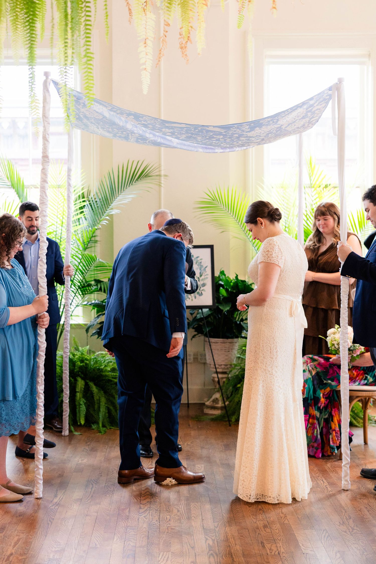 Bride and groom bow heads during ring exchange under decorated chuppah at indoor Jewish wedding ceremony.
