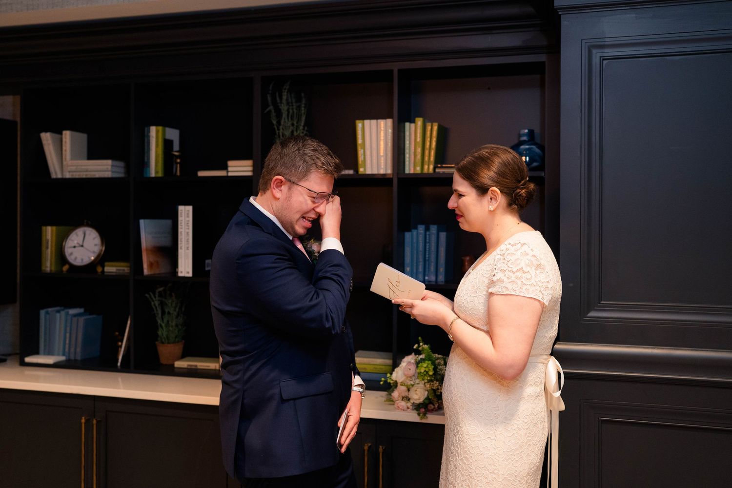 Groom emotionally covering face while bride reads heartfelt vows in dark elegant library setting.