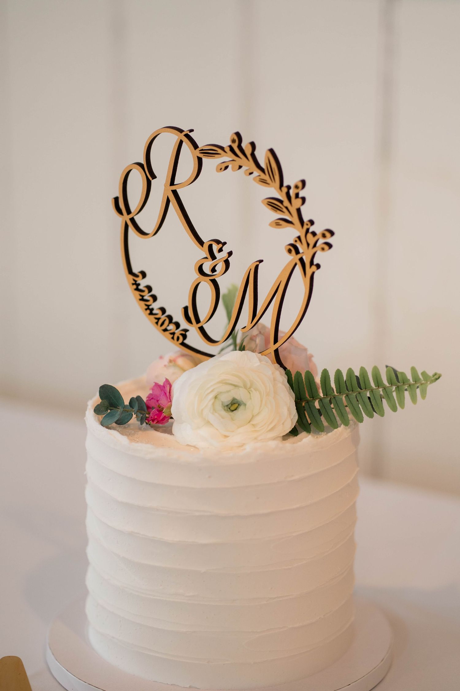 Close-up of white textured wedding cake with laser-cut wooden monogram topper featuring R&M initials and floral decor.