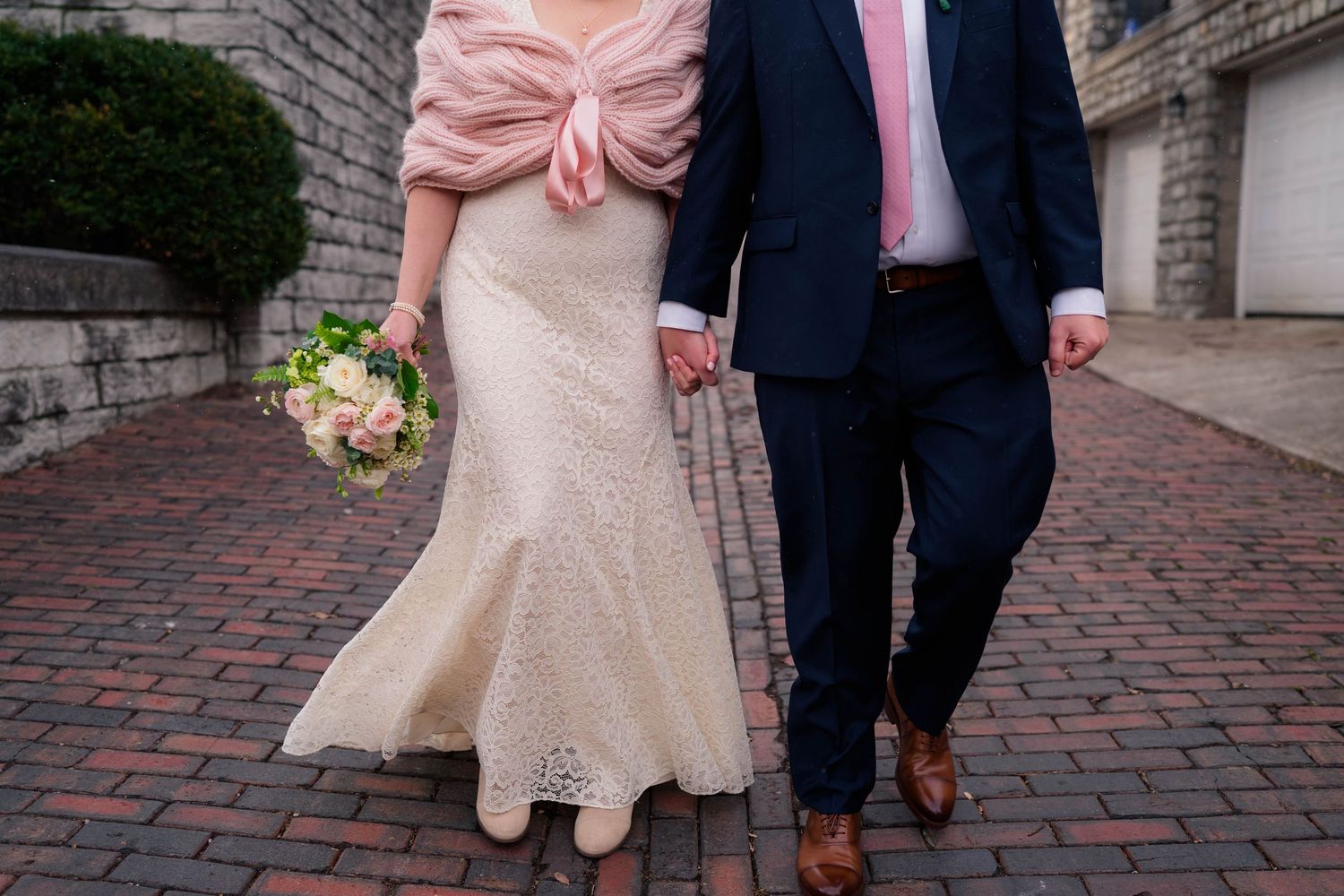 Close-up of bride in lace wedding gown with pink wrap and groom in navy suit holding hands on brick pavement.