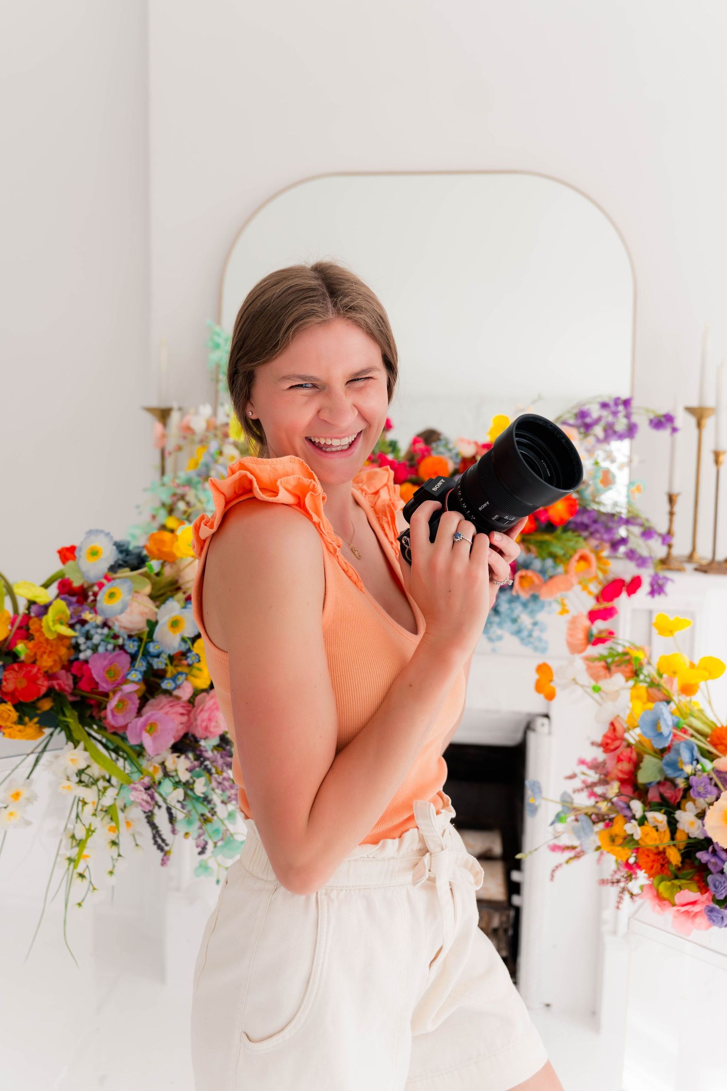 Smiling woman holding a camera in an orange top and white shorts, standing in front of colorful floral arrangements.