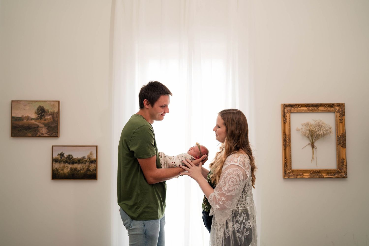 Young couple sharing tender moment with newborn baby in bright gallery room with framed artwork on white walls.
