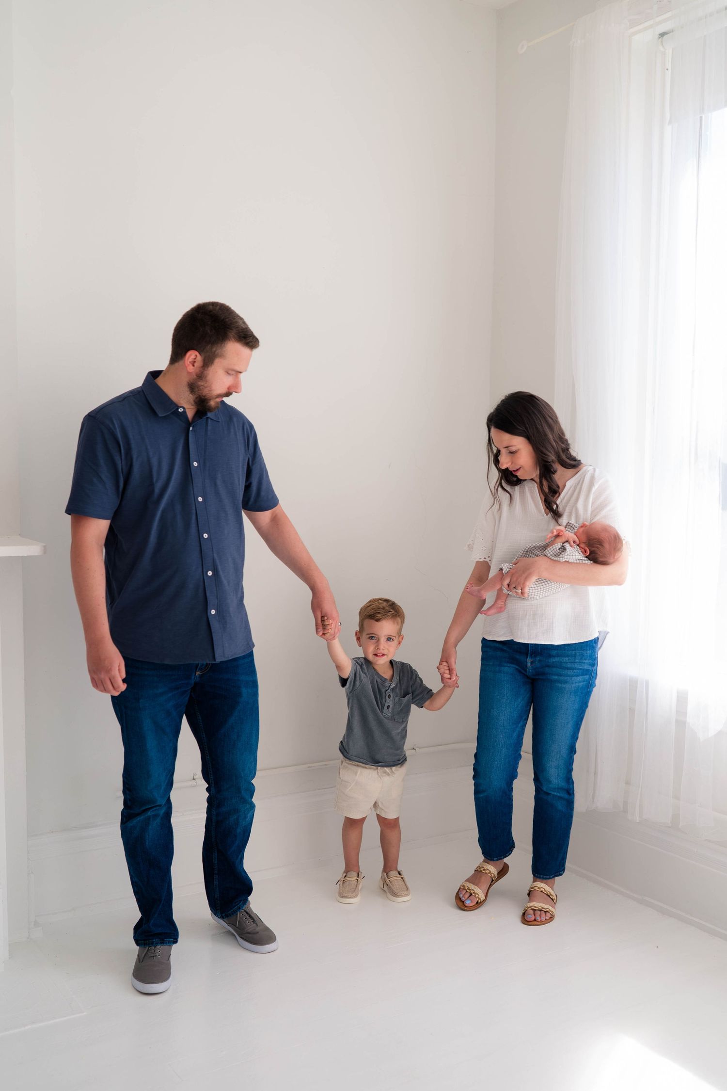 A young family of four poses in a bright white room, parents holding hands with toddler son while mom cradles newborn baby.